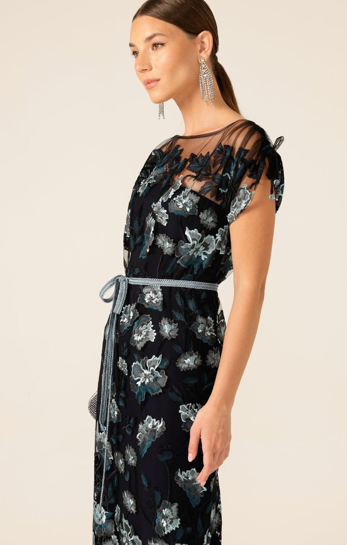 A woman in the SACHA DRAKE LOVE POTION MAXI IN TEAL AQUA, a boat-neck maxi with blue and white embroidered floral mesh, sheer sleeves, a teal aqua waist tie, and statement earrings stands against a neutral background.